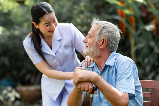 Caring nurse helping senior man sitting on bench in gaden. Asian woman, caucasian man. Happy smile.